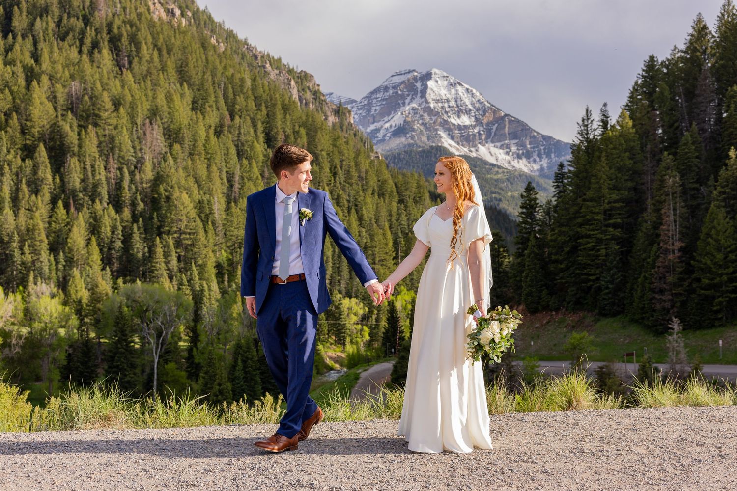 Wedding couple shares a moment on mountain path with evergreen forest and peaks in background.