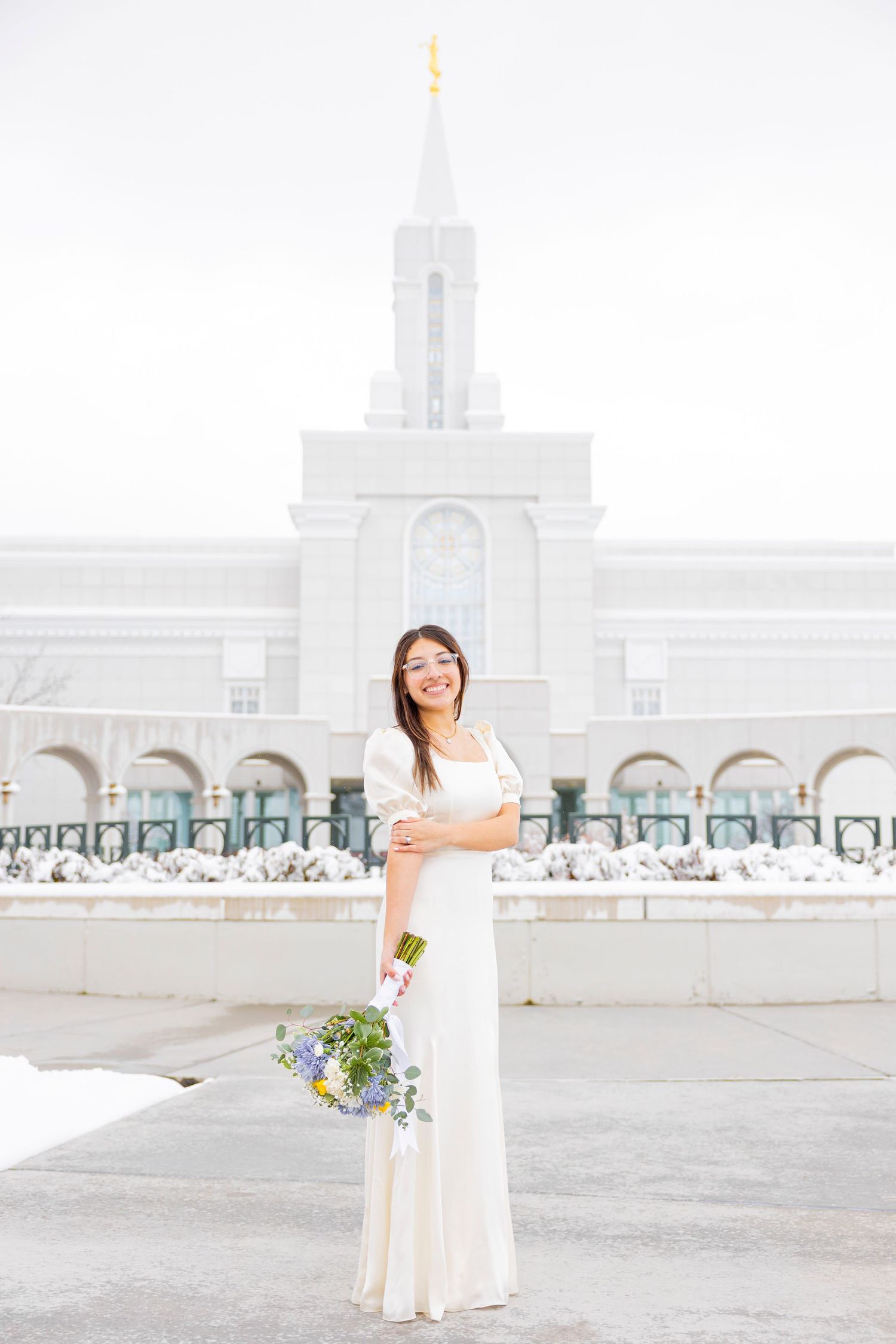 A bride in a white dress poses with a bouquet against a light-colored architectural backdrop.