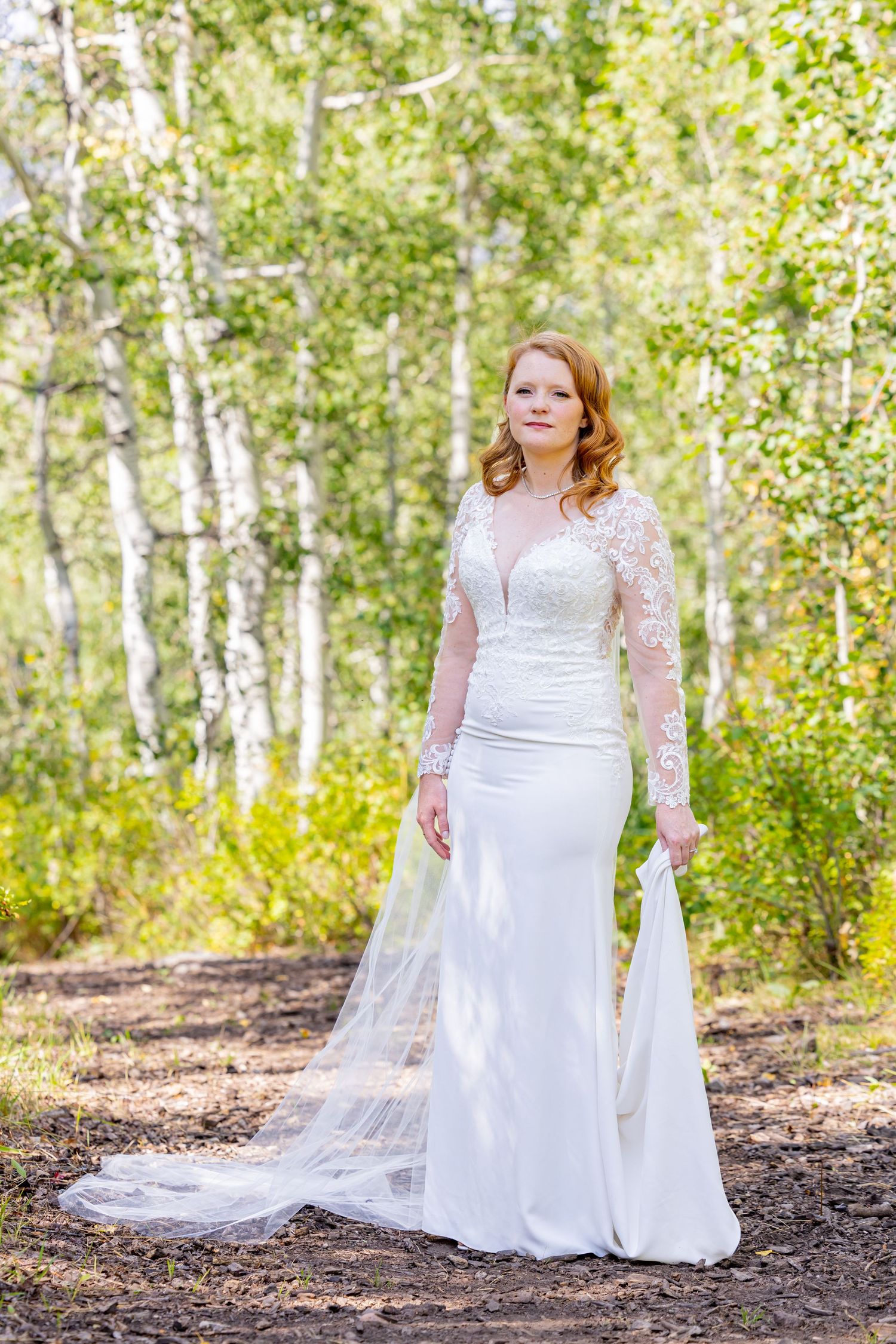 A bride in a long-sleeved lace wedding gown poses in a sunlit birch forest.