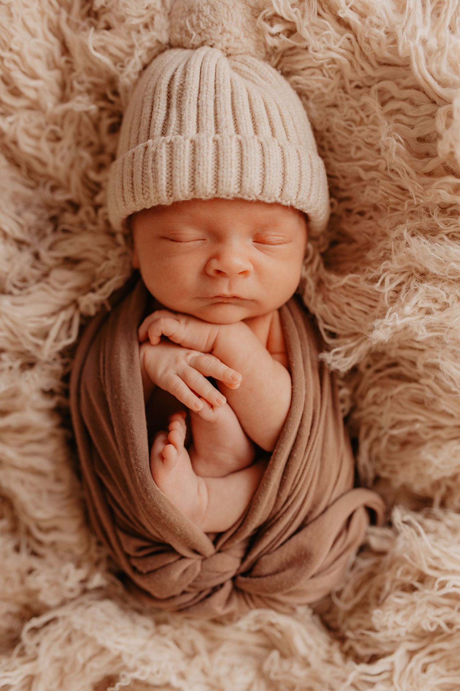 Newborn baby wearing knit hat lies peacefully on soft furry blanket in neutral tones.