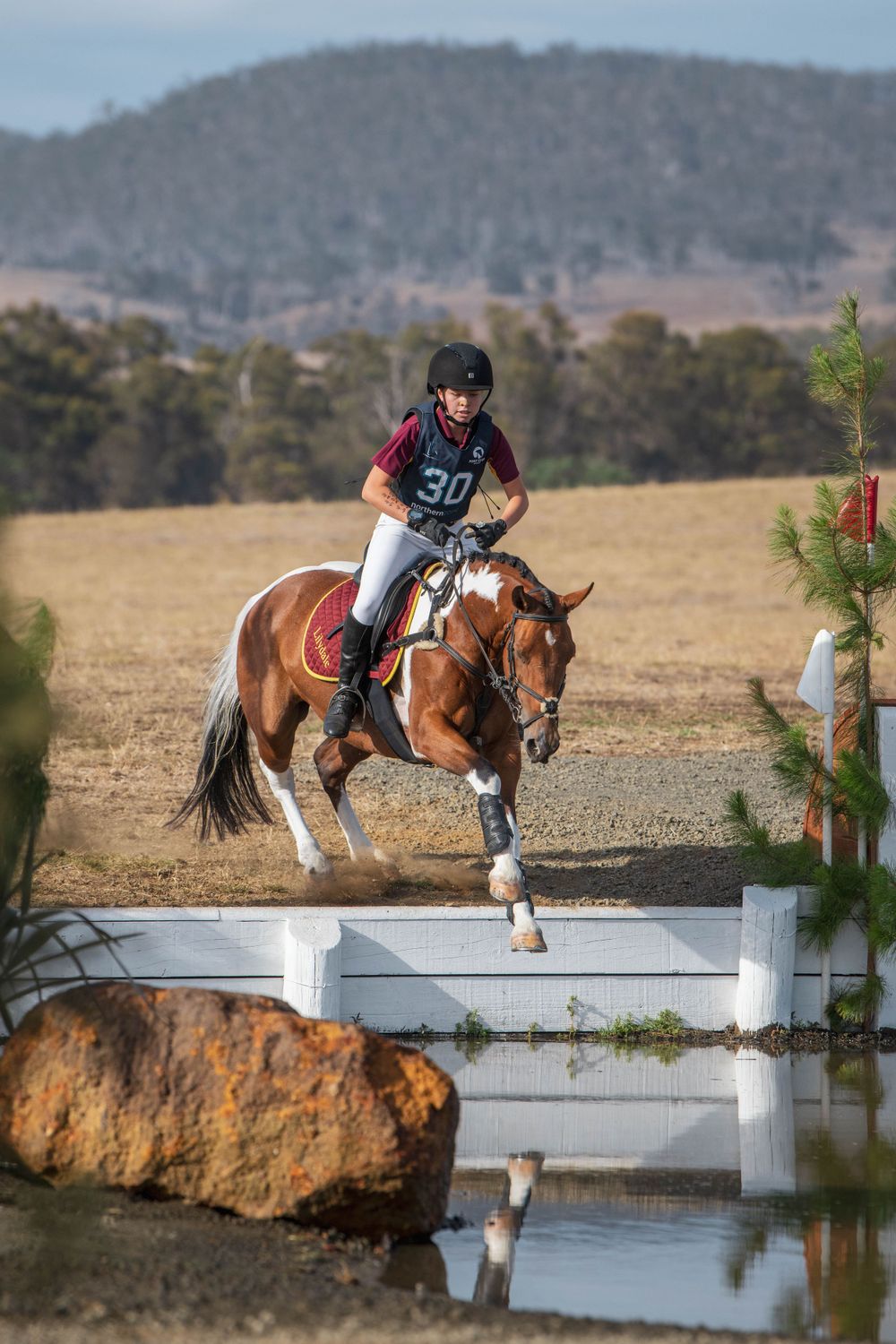 A rider jumps their chestnut horse over a white water obstacle during an equestrian event.