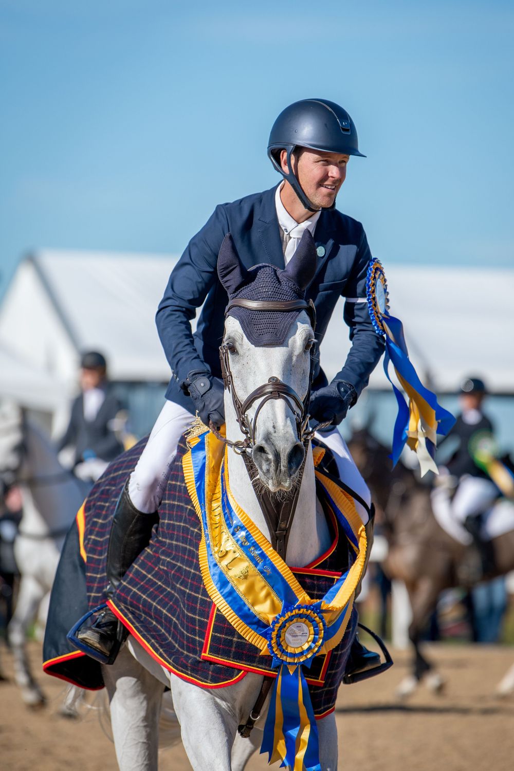 Smiling equestrian rider in navy jacket on decorated white horse with blue and gold ribbons at competition.