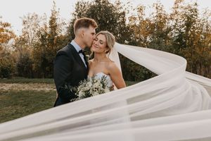 A wedding couple embraces in a field at sunset while the bride's long veil flows dramatically in the wind.