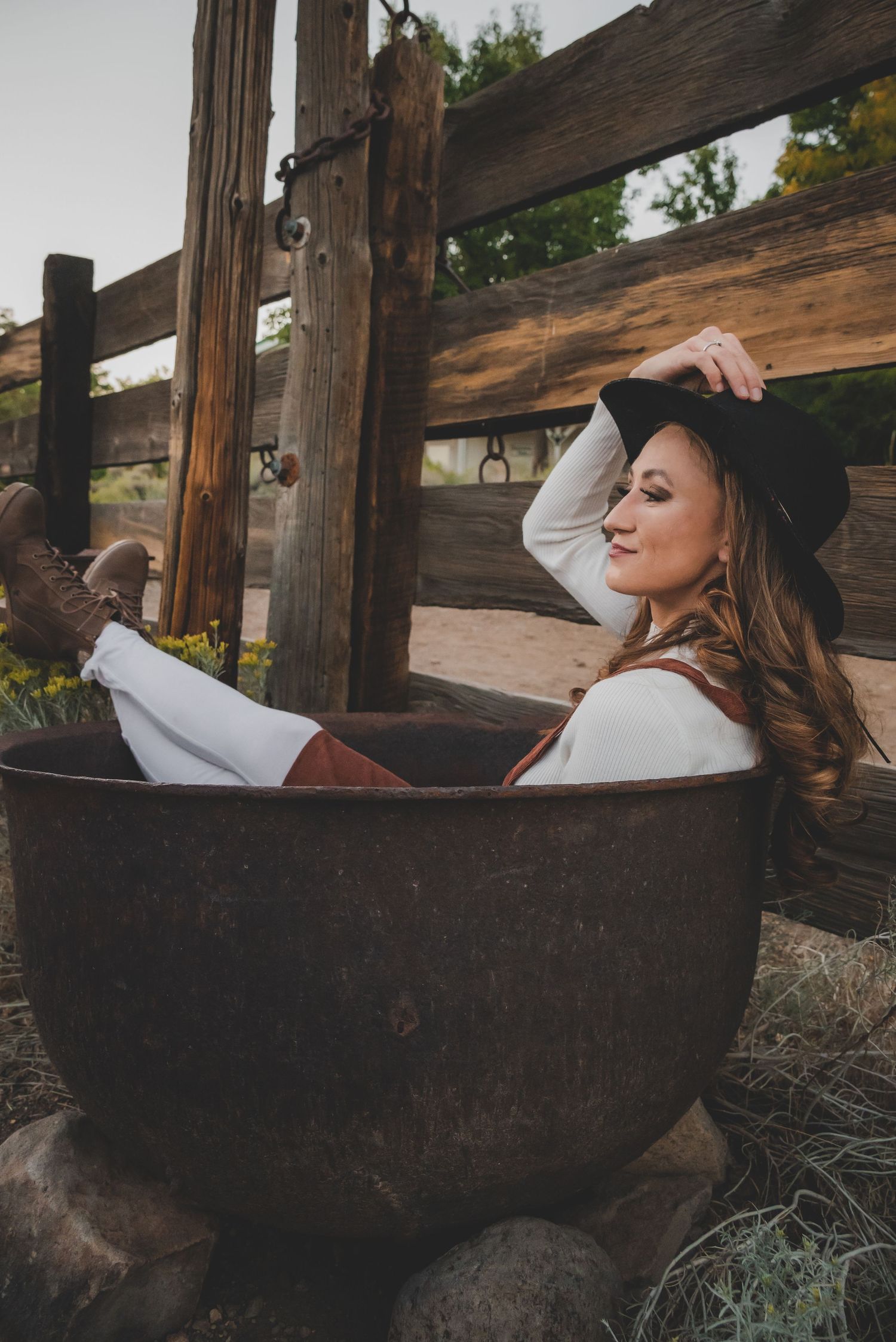 Smiling woman in cowboy hat relaxes in rustic metal tub on farm.