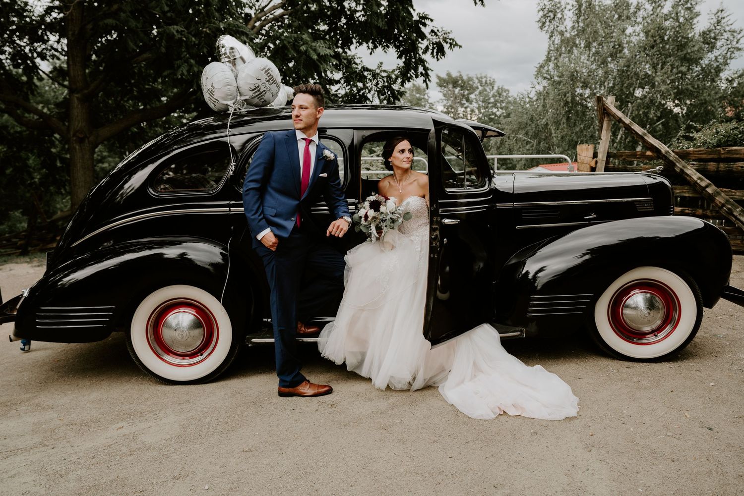 bride sitting in classic car with whitewall tires. Groom leaning on car.