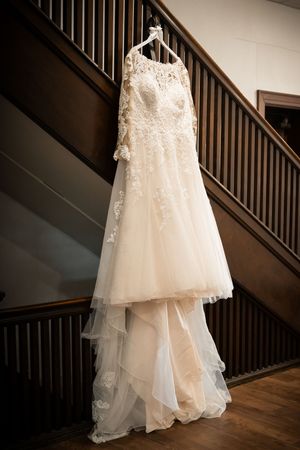 Elegant lace wedding gown with long sleeves hanging on a hanger against a wooden staircase railing.