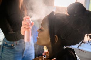 A makeup artist sprays setting mist on a young woman with dark hair in a bun during a beauty session.