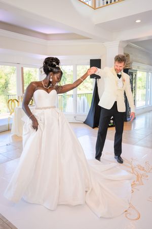 Bride and groom share first dance, bride in strapless white gown, groom in white tuxedo jacket, elegant venue.