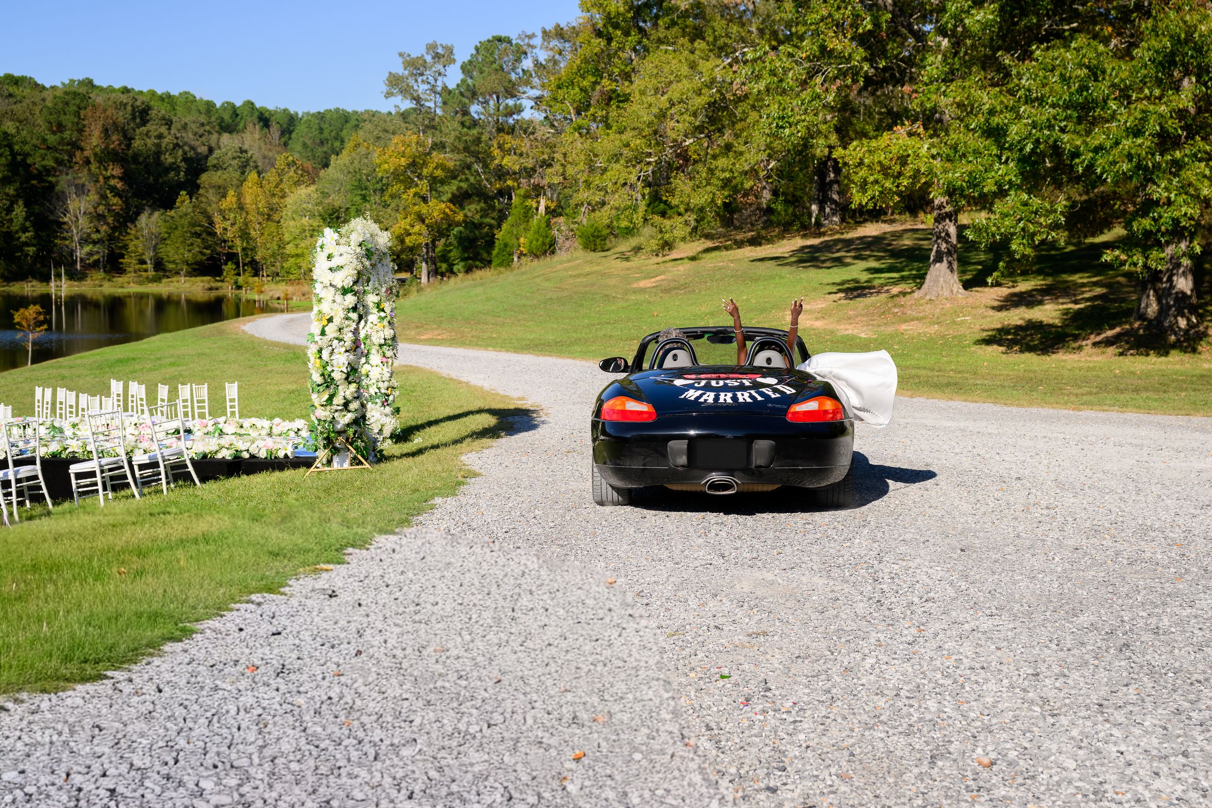 Black sports car driving down gravel path at outdoor wedding venue with white floral arch and chairs.