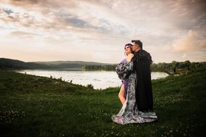 Couple embracing in flowing gown and black cloak on green hillside with scenic river and cloudy sky.