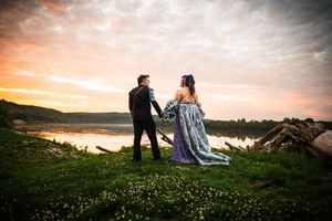 Couple in formal attire standing on grassy lakeside at sunset, bride in blue gown holding groom's hand.