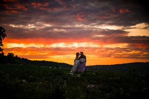 Couple embracing at sunset with dramatic orange and red sky over rolling green hills.