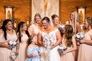 Bride in white gown laughs with bridesmaids in champagne dresses holding floral bouquets inside a church.
