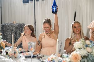Bridesmaids in blush dresses celebrate at wedding reception, one raising a blue bottle in excitement.