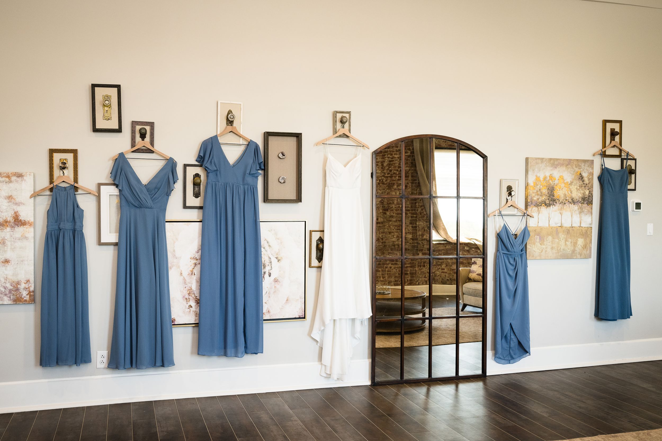Blue bridesmaid dresses displayed on hangers against a white wall with framed photos.