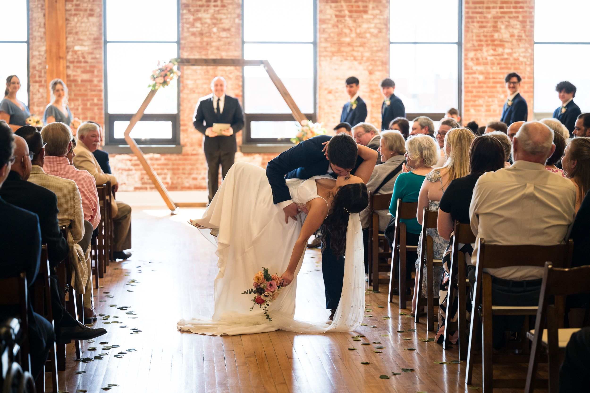 Wedding ceremony in a rustic brick venue with guests watching the couple's first kiss.