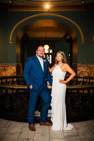 Bride in white gown and groom in blue suit pose elegantly inside a grand historic venue with arched decor.