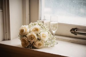 White rose bridal bouquet with baby's breath beside two champagne flutes on a window sill.