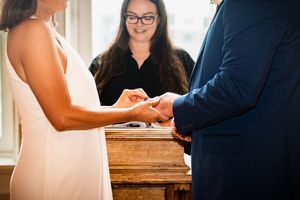 Couple exchanging rings during wedding ceremony with smiling officiant in glasses standing behind wooden altar.