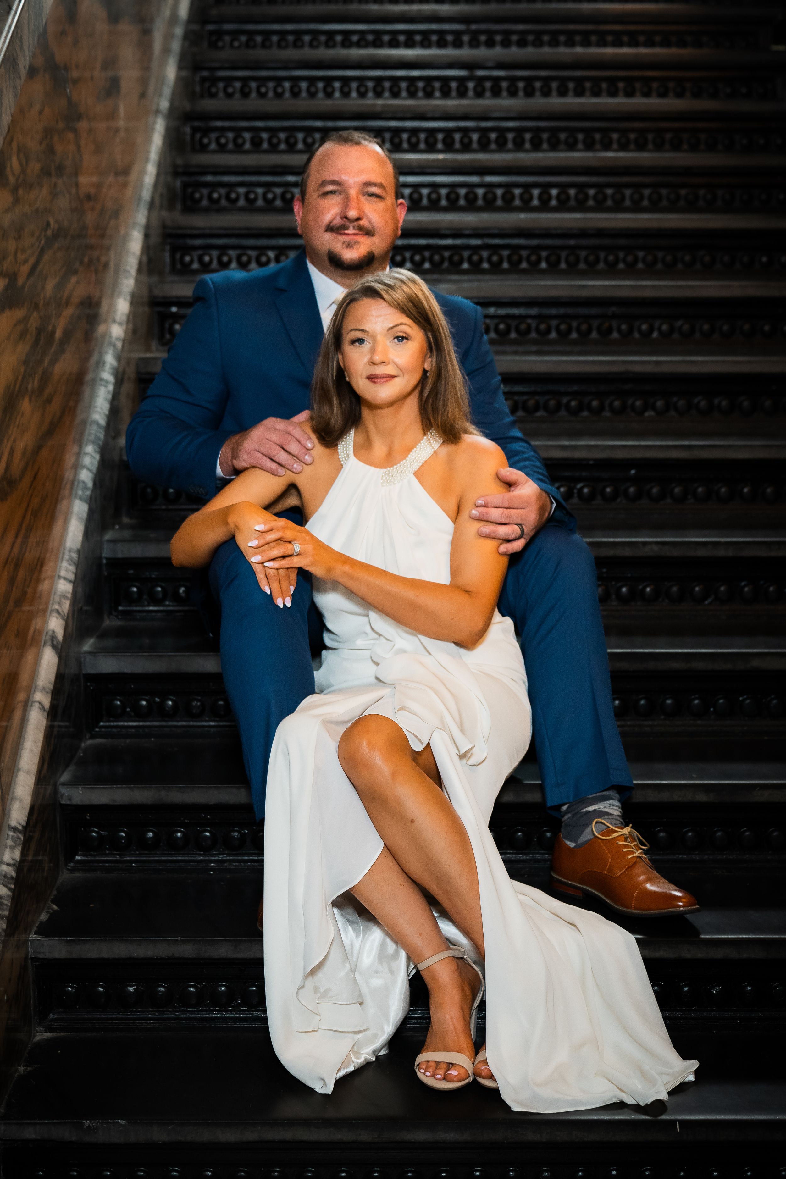 Couple in formal attire poses on dark staircase, man in blue suit embraces woman in white halter dress.