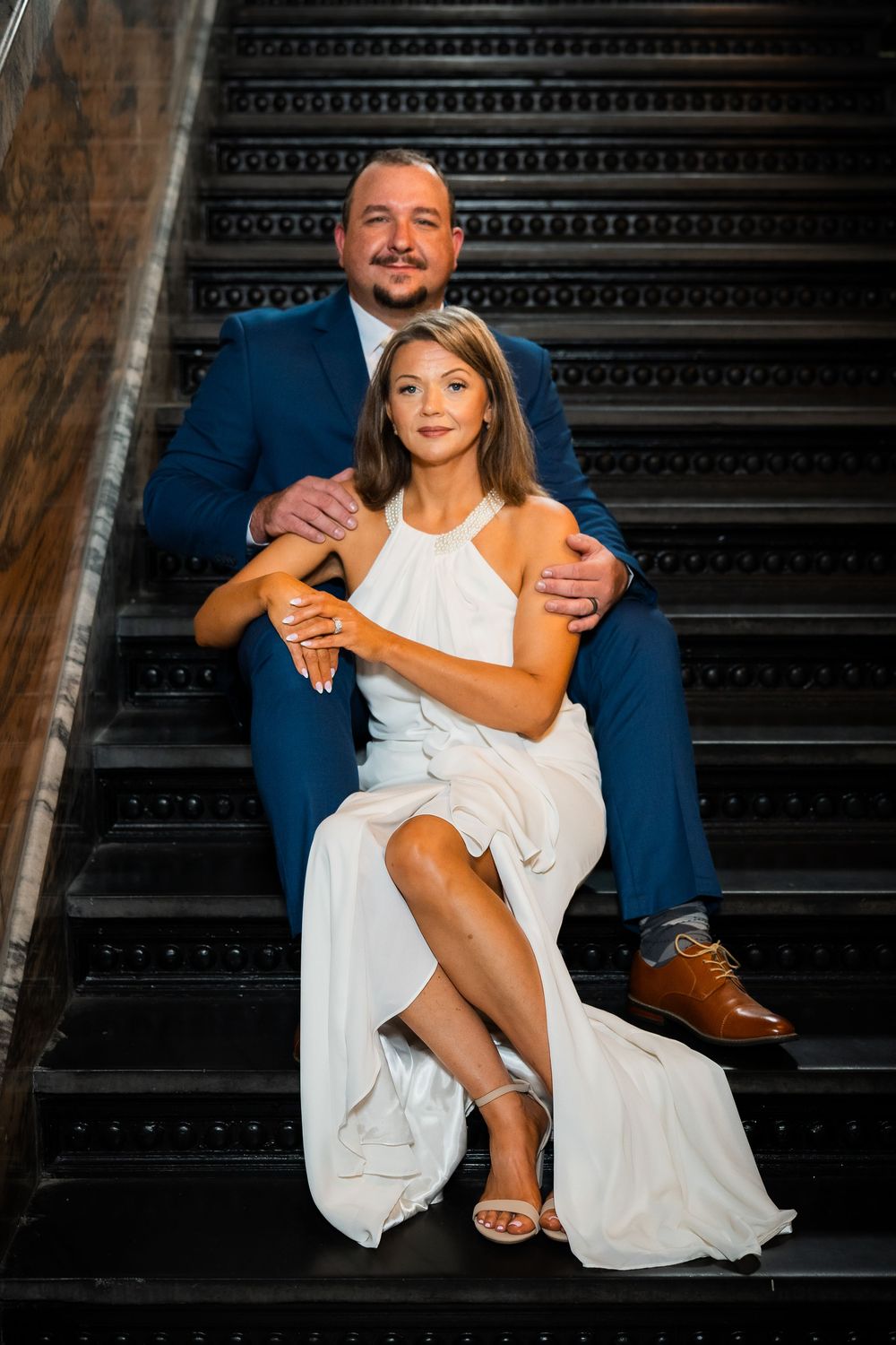 Couple in formal attire poses on dark staircase, man in blue suit embraces woman in white halter dress.