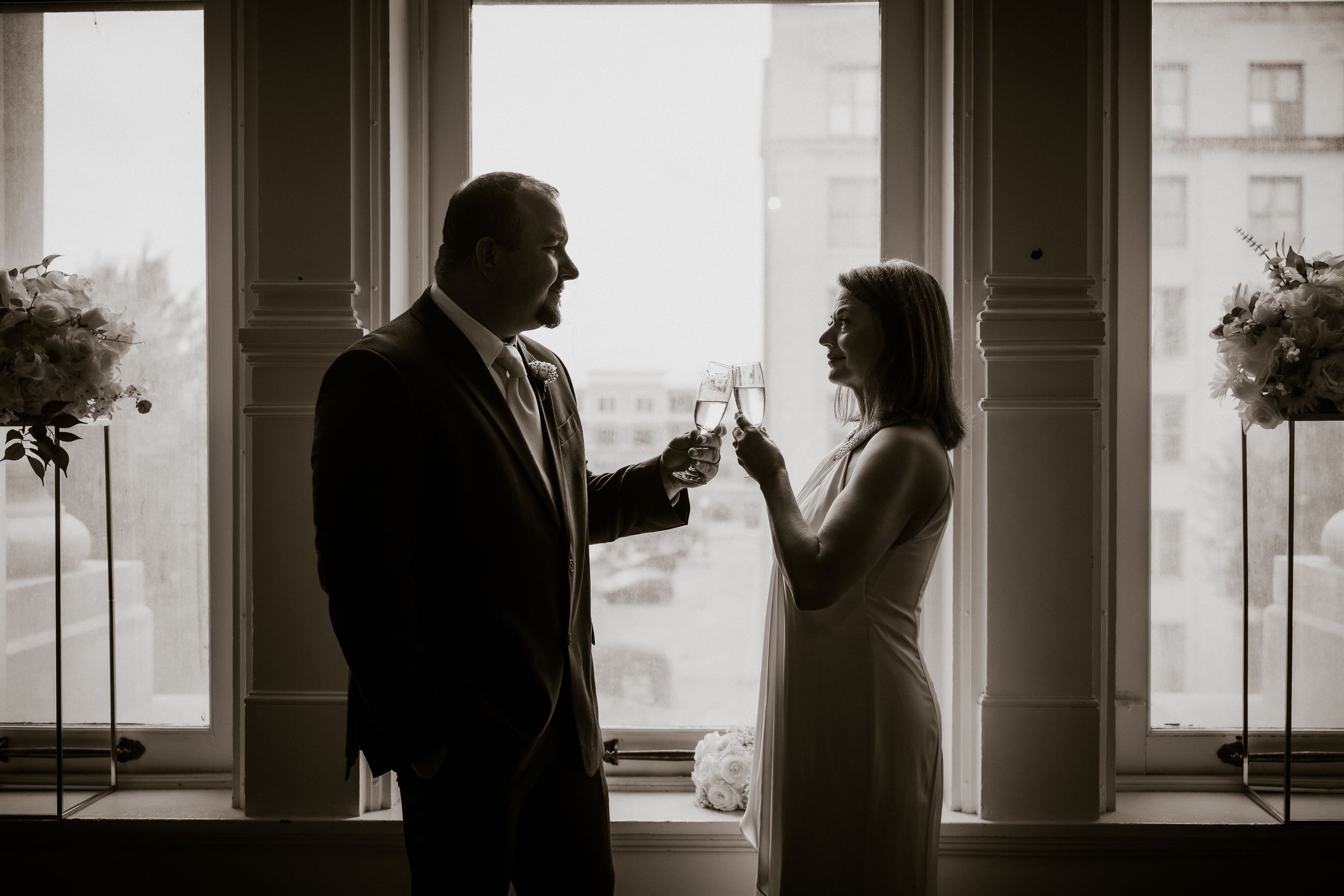 Black and white photo of a couple toasting with champagne glasses by a window, celebrating a special occasion.