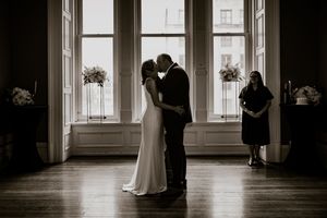 Black and white photo of a couple sharing their first kiss at an indoor wedding ceremony with large windows.
