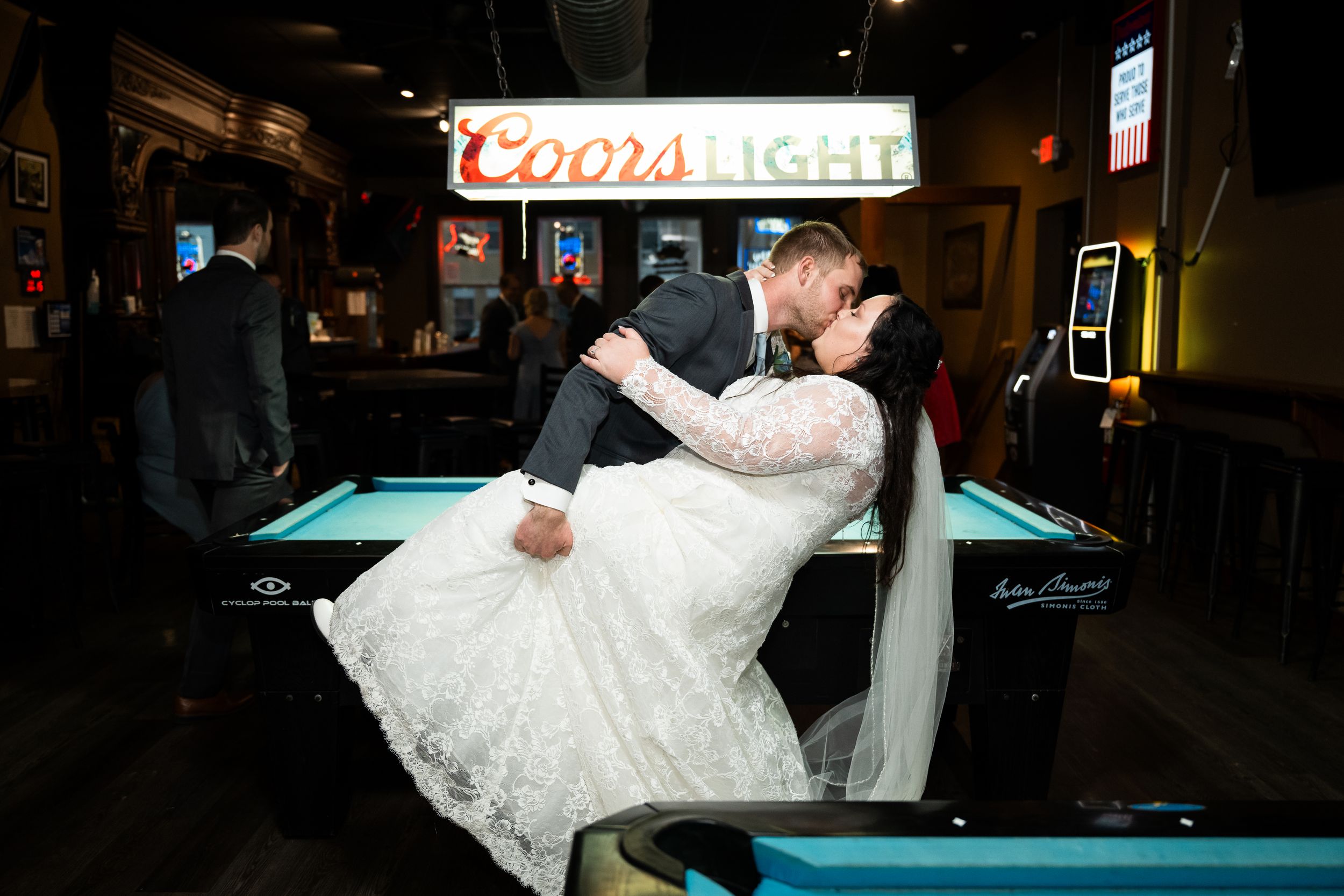 A couple in wedding attire shares a romantic dip pose in front of a pool table at a bar with a Coors Light sign glowing above.
