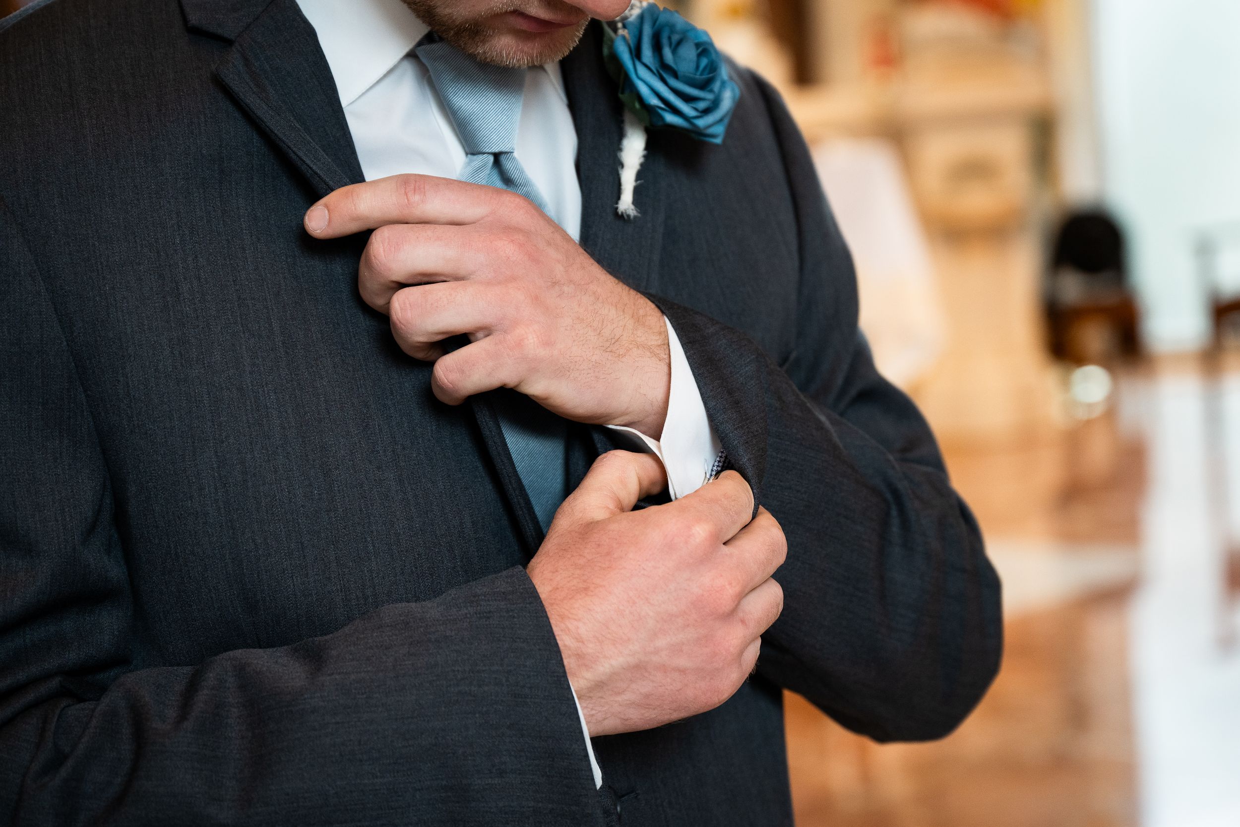 A groom in a dark suit and blue tie adjusts his sleeve cuff while wearing a teal boutonniere.