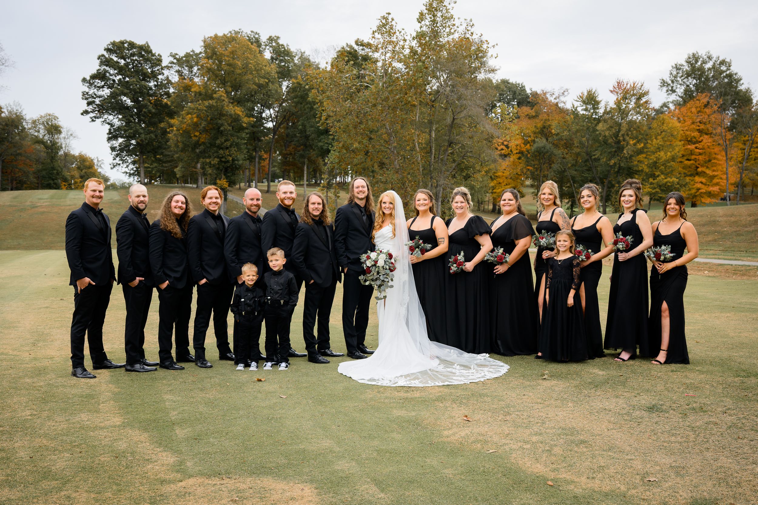 Wedding party in black attire poses outdoors on golf course with autumn trees, bride in white gown at center.
