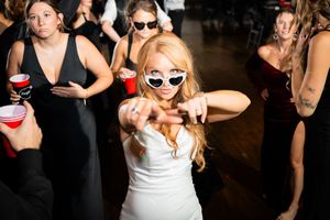 Bride in white dress and heart sunglasses dancing at wedding reception with guests in black dresses holding red cups.