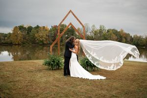 Bride and groom kissing under a geometric wooden arch by a lake, with veil flowing in the wind on an autumn day.