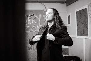 Black and white photo of a long-haired man in a dark blazer adjusting his jacket in a graffiti-covered room.