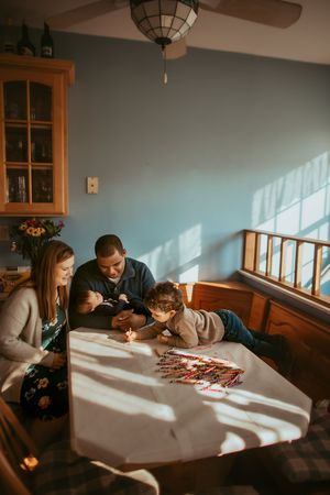 A group gathers around a sunlit table to work on a puzzle together in a cozy room with wooden windows.