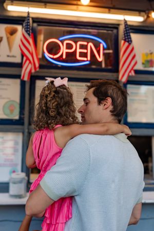 A pink-dressed child being held at an American diner with neon OPEN sign and US flags in the background.