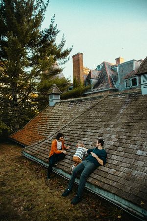 Two people relax on an old wooden shingled rooftop at sunset with pine trees in the background.