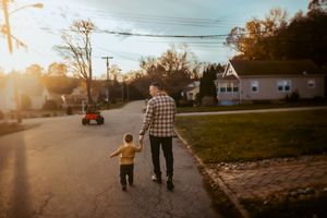 A silhouetted figure and child walk down a suburban street at sunset with golden sunlight streaming through trees.