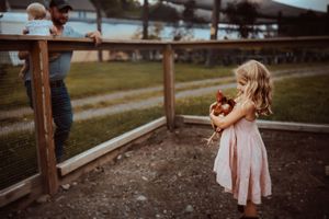 A toddler in a pink dress walks along a rustic wooden fence in a pastoral outdoor setting.