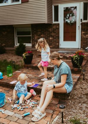 Children playing with toys and crafts on a brick patio outside a house with colorful flowers in the background.