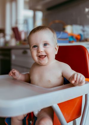 Happy baby sitting in orange high chair with white tray showing playful expressions during mealtime.