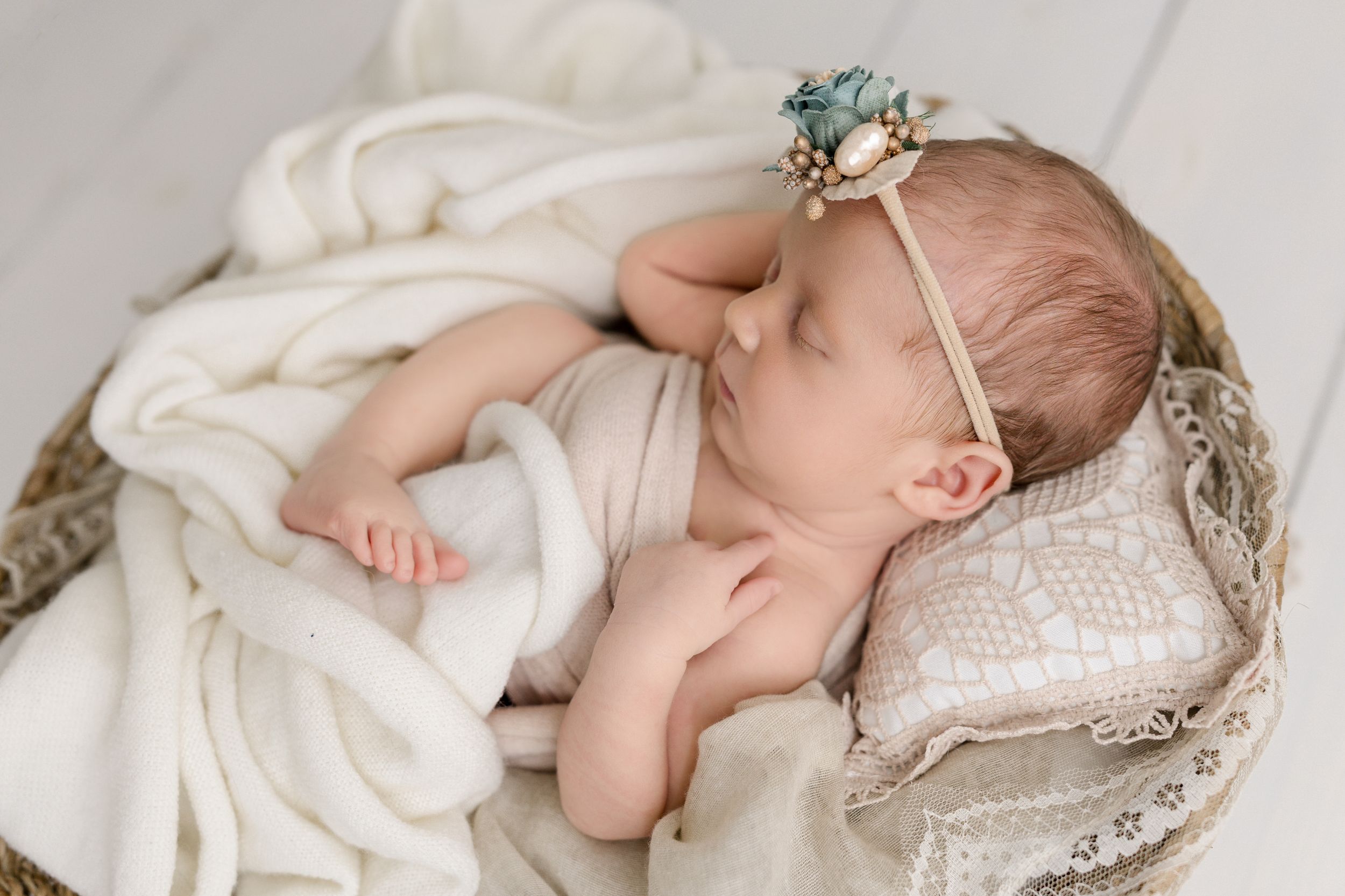 Newborn baby sleeping peacefully on white lace pillow wearing delicate floral headband and wrapped in soft blanket.