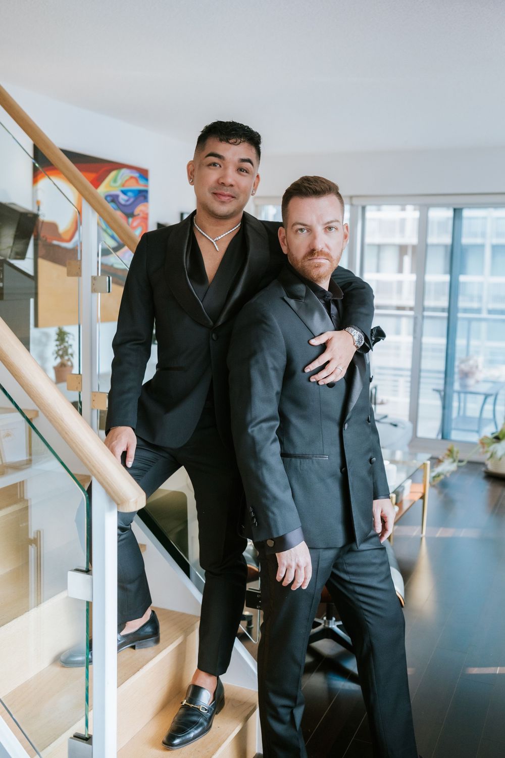Two people in dark formal suits pose together on modern wooden stairs in a bright interior space.