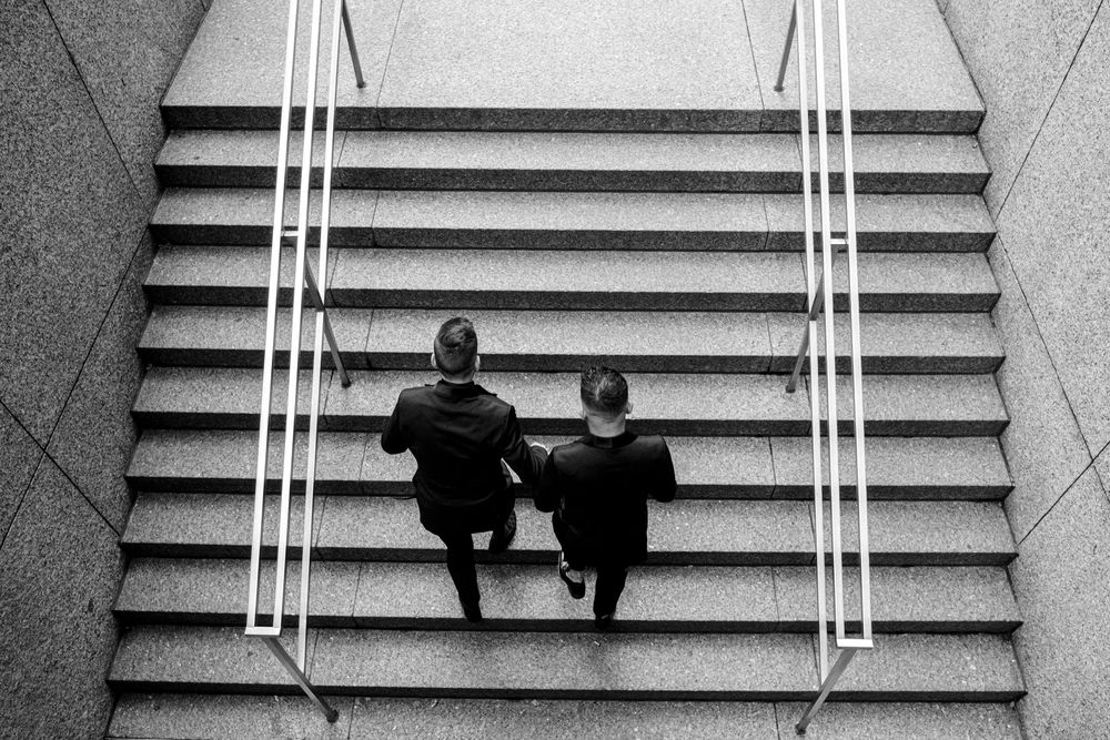 Two figures sit together on wide concrete steps between white handrails in a black and white architectural photograph.