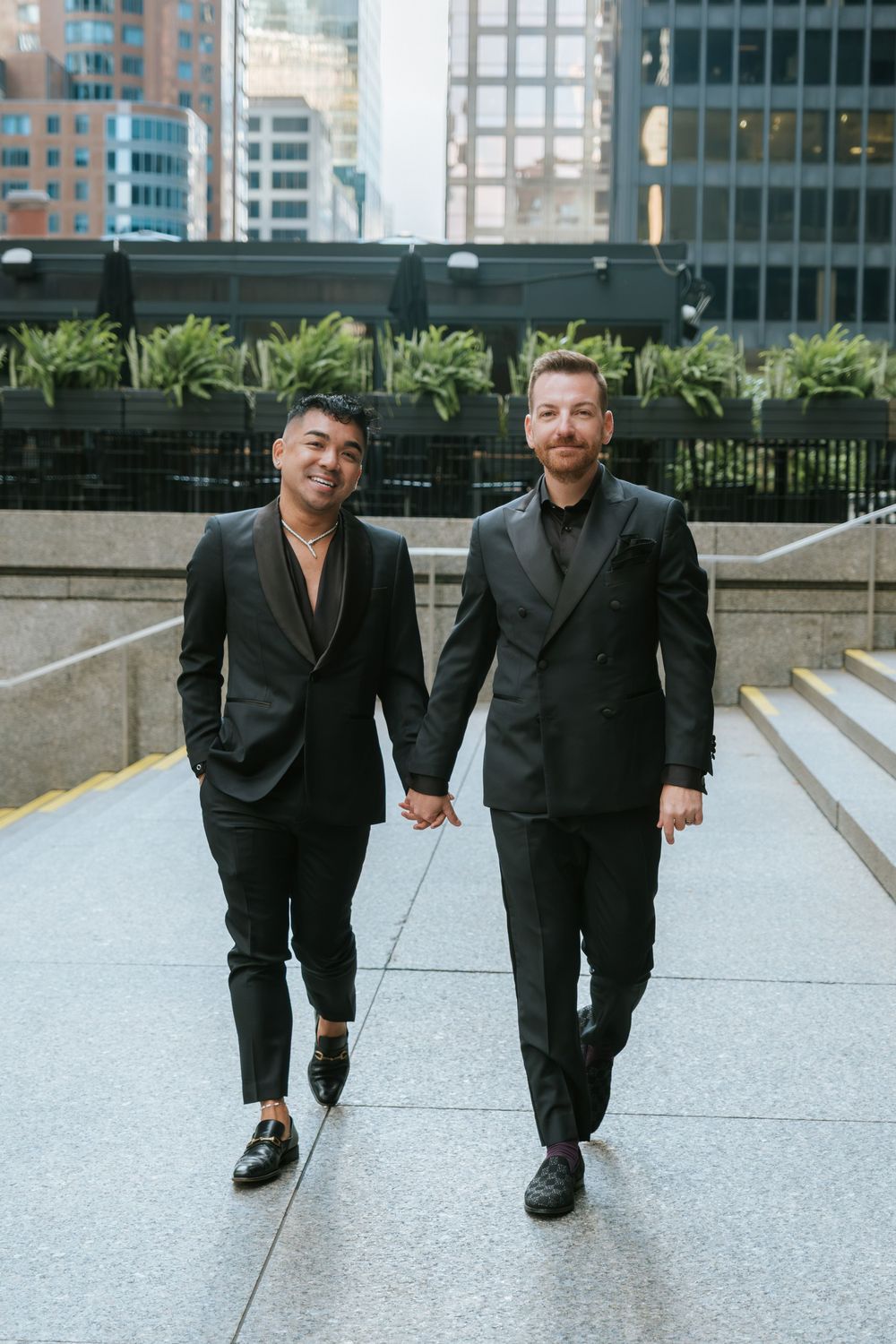 Two people in black suits walk together on concrete steps with urban buildings visible in the background.