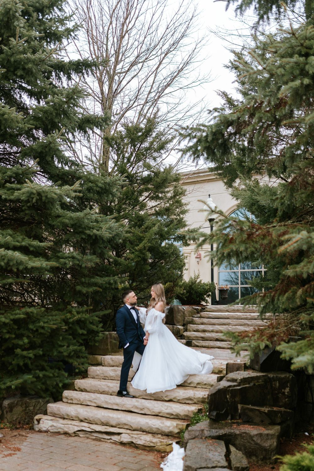 A couple sits together on rustic stone steps surrounded by evergreen trees in a romantic outdoor setting.