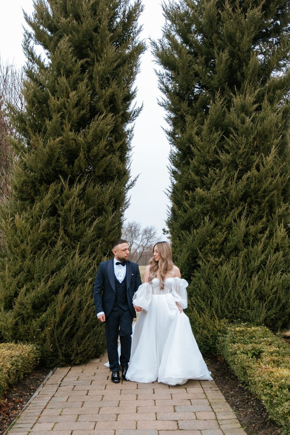 A couple in wedding attire walking along a stone path between tall evergreen trees in a garden setting.