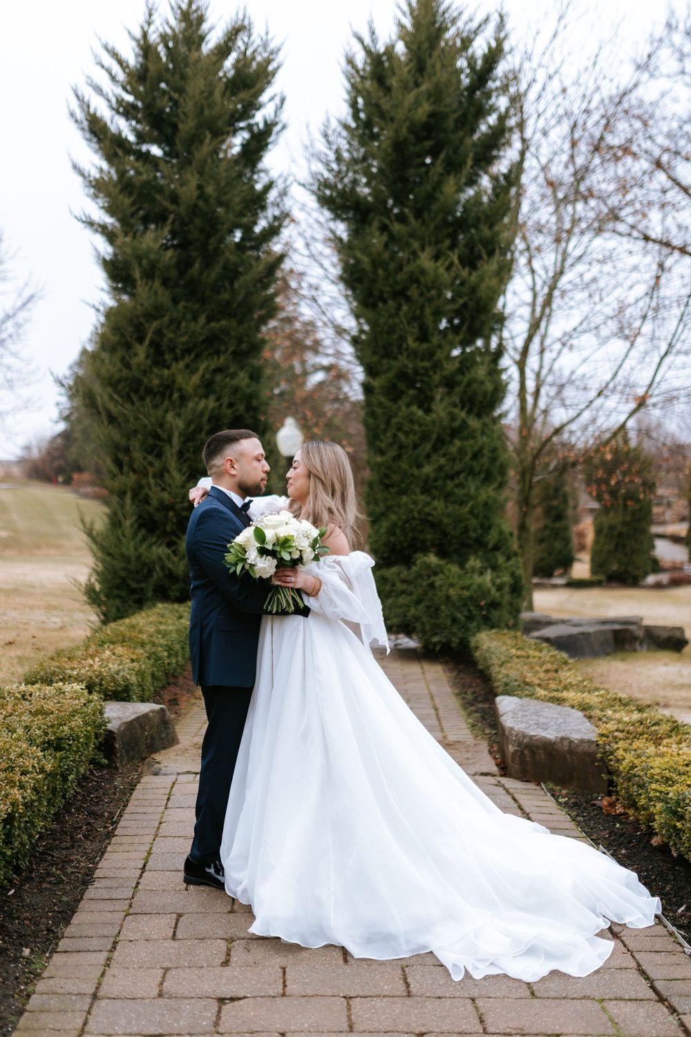 A romantic wedding couple shares intimate moments on a stone pathway surrounded by evergreen trees in winter.