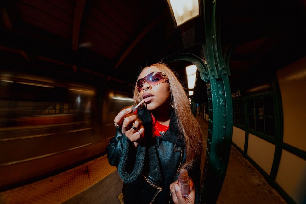Woman applies lipstick in subway station with motion-blurred train behind her.