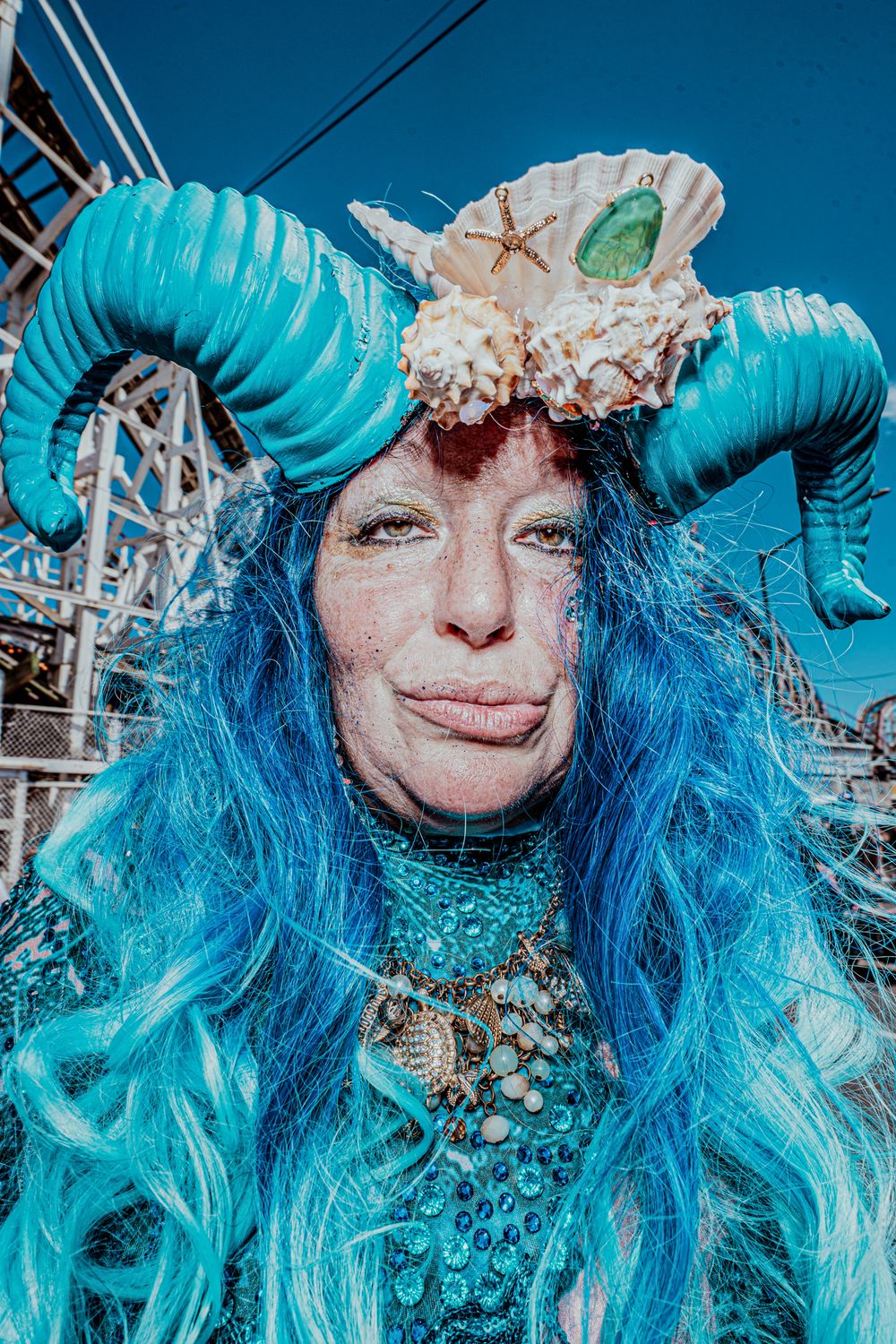 Woman in elaborate teal mermaid costume with blue hair and shell horns at Coney Island parade.