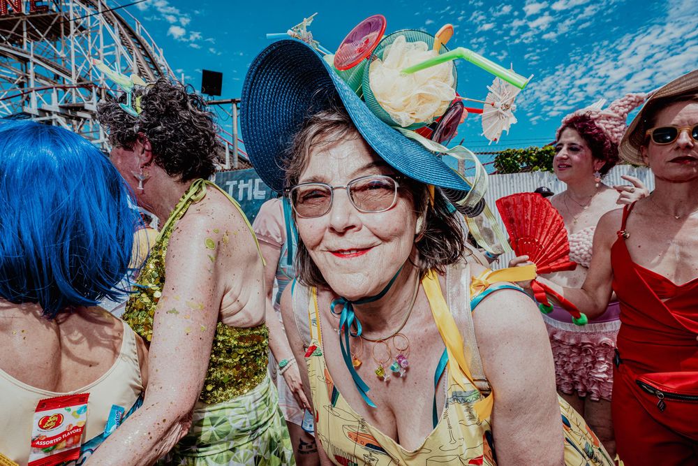 Woman in colorful dress and blue hat smiles at vibrant outdoor festival.